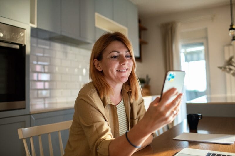 Kvinna sitter vid köksbord och tittar på sin mobiltelefon.