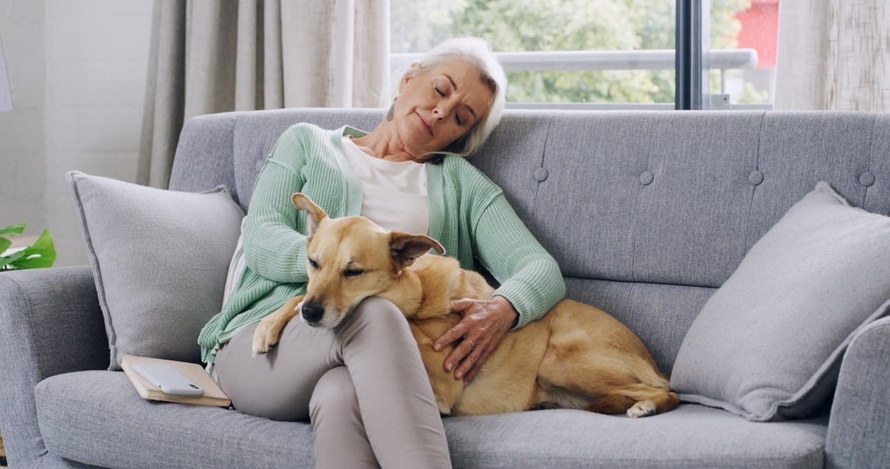 Kvinna sitter i soffa och blundar med en hund vilandes i knät.