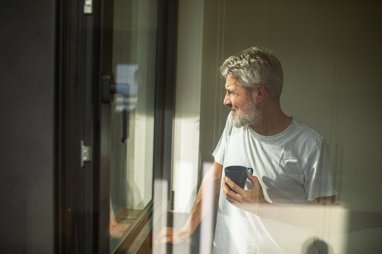 Man står med kaffemugg i handen och tittar ut genom fönster.